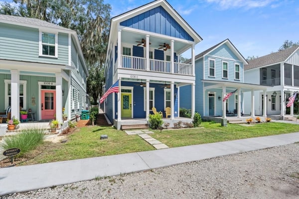 Row of colorful modern cottage-style homes with front porches, white railings, and American flags along a tree-lined street.