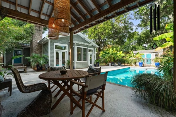 Covered patio with dining table overlooking swimming pool and modern cottage with green shutters