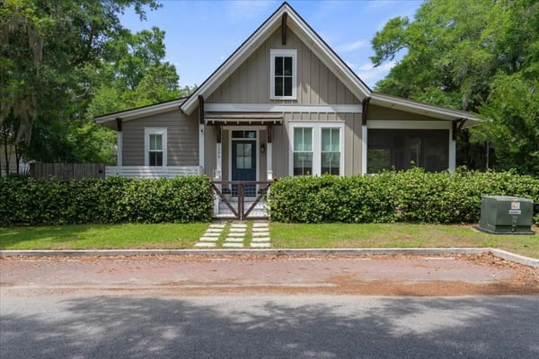 Lime-green cottage with white trim, covered porch, ceiling fan, and attached garage surrounded by trees