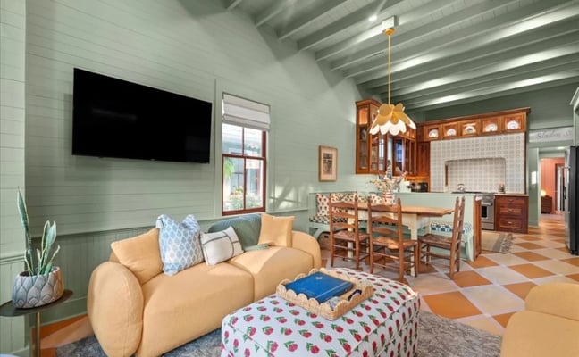 White cottage with green front door, covered porch, and white picket fence surrounded by oak trees and tropical plants